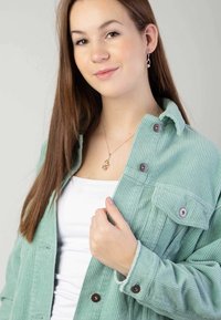 Mintgrüne Kordjacke mit Kragen, Knopfverschluss und einer Brusttasche. Das Model trägt ein weißes Oberteil und silbernen Schmuck.