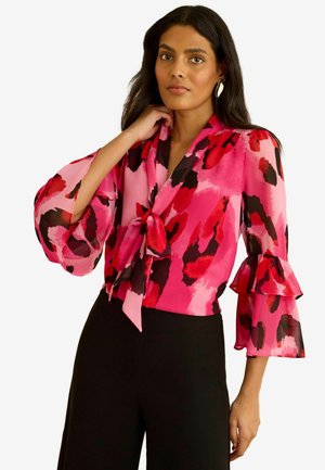 Pink blouse with black and red abstract pattern, tie neckline, ruffled sleeves, and lightweight fabric paired with black bottoms.