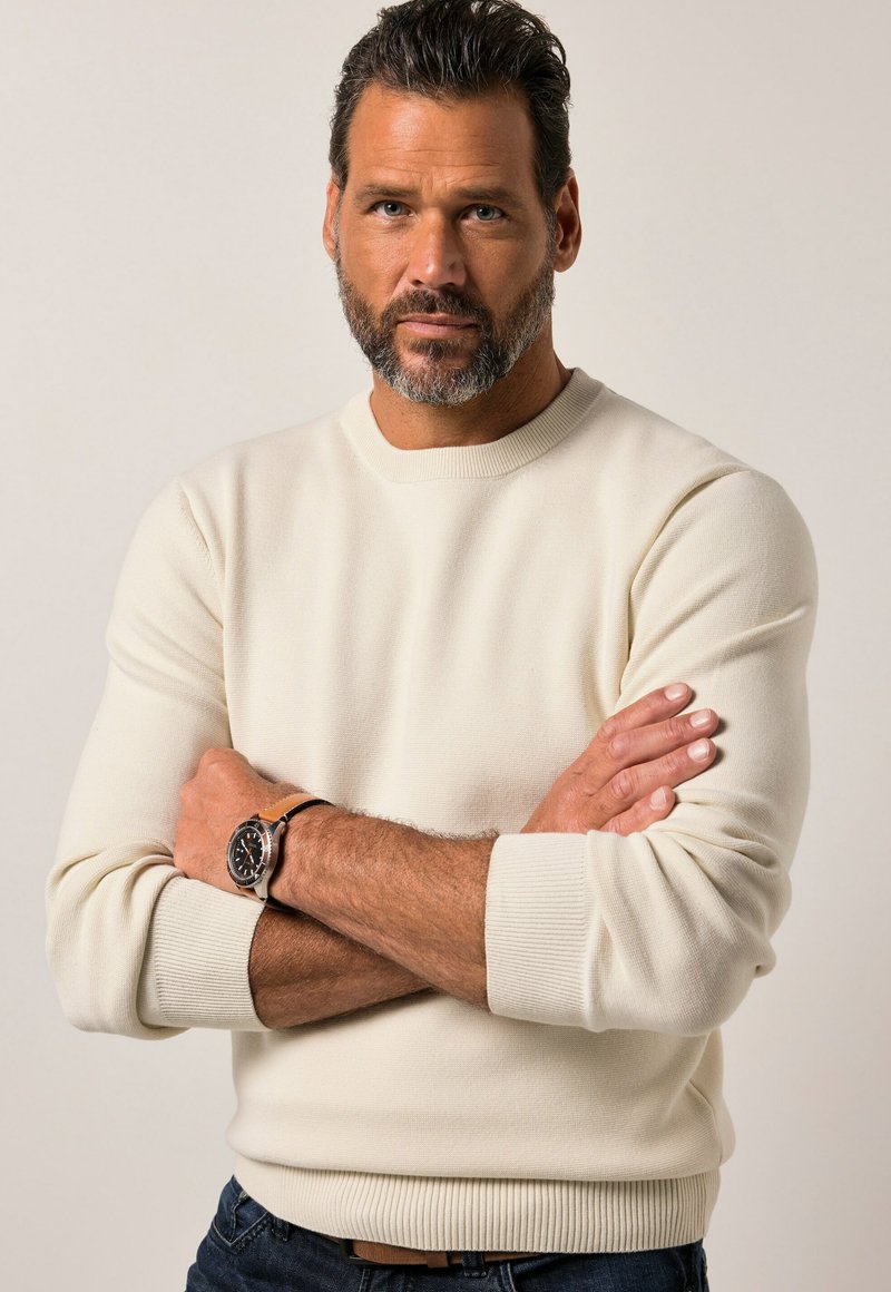Mann mit Bart, der einen cremefarbenen Pullover und eine Armbanduhr trägt, steht selbstbewusst mit verschränkten Armen vor einem neutralen Hintergrund.