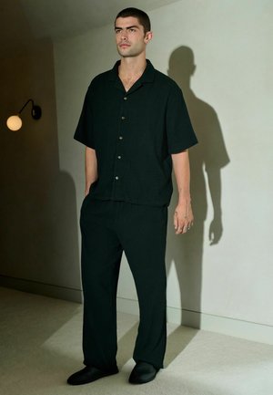 Czarna koszula z krótkim rękawem i guzikami, dopasowane luźne spodnie oraz czarne buty wsuwane, na tle neutralnego tła i miękkiego oświetlenia.