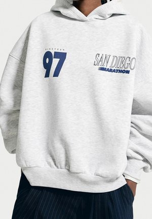 Hellgrauer Hoodie mit blauem Aufdruck „Nineteen 97“ und „San Diego Marathon“, kombiniert mit dunkler Nadelstreifenhose.