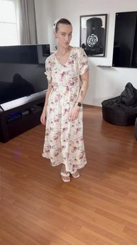 Pessoa com vestido midi floral e sandálias brancas a estar em chão de madeira numa sala com televisão, pufe e obras de arte emolduradas nas paredes.