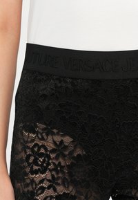 Μαύρο δαντελωτό σορτσάκι με floral σχέδιο, ελαστική μέση με ανάγλυφο κείμενο "FUTURE VERSACE JEANS" και ημιδιάφαστη υφή.