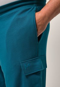Teal cargobyxor med en mjuk textur, som har en sidoficka och en broderad logodetalj på ficklocket.