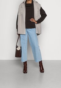 Světle šedá bezrukávová vesta s kapsami, tmavě hnědý top, světle modré džíny a hnědé ankle boots s krokodýlím vzorem. Drží hnědou tašku.