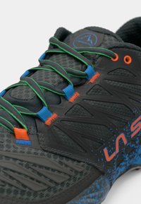 Szürke sportcipő, hálós felsőrésszel, élénk zöld, kék és narancssárga cipőfűzőkkel, valamint texturált részletekkel ellátott kék pöttyös talppal.