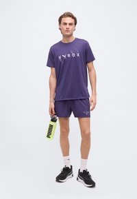 Άνδρας που φοράει μοβ αθλητικά ρούχα HYROX PUMA κρατώντας ένα κίτρινο παγούρι PUMA, στέκεται με μαύρα και λευκά αθλητικά παπούτσια και λευκές κάλτσες.