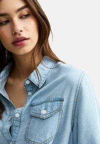 Camicia in denim azzurro chiaro con colletto a bottoni, taschino sul petto e cuciture a contrasto. Texture liscia e vestibilità rilassata.