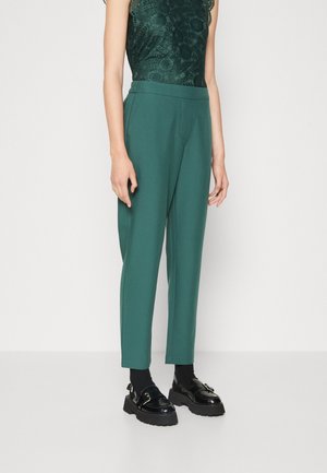 Personne portant un pantalon vert taille haute et fuselé, des chaussettes noires, des mocassins noirs brillants avec boucle et un haut sans manches en dentelle vert foncé.