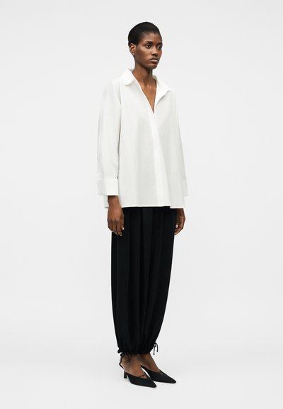 Žena stojící, oblečená v nadměrně velké bílé košili s dlouhým rukávem, volných černých kalhotách se stahovací šňůrkou a černých lodičkách s špičatou špičkou na jednobarevném pozadí.