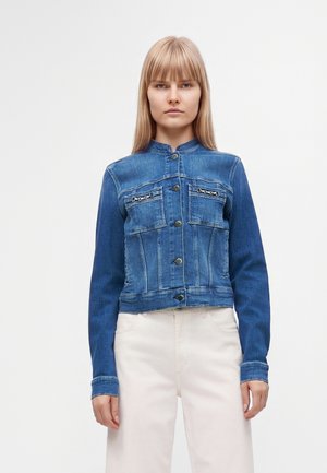 Femme portant une veste en jean bleue à fermeture par boutons et deux poches poitrine avant, associée à un pantalon clair sur un fond uni.