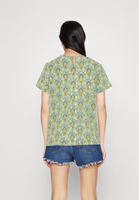 T-shirt à manches courtes avec un motif floral vert, bleu et beige ; fermeture à boutons au dos ; porté par-dessus un short en denim avec des ourlets effilochés.