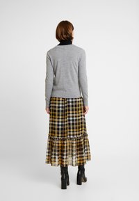 Grå tröja med en figursydd design, bärs över en lång kjol i gult och svart rutmönster med en volangkant och lager-på-lager-textur. Svarta ankelstövlar.