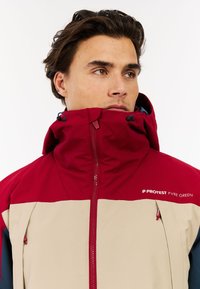 Rote und beige Zip-Jacke mit hohem Kragen, Kordelzug und Logodetail auf der Brust; aus glattem, strukturiertem Material gefertigt.
