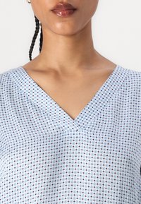 Blusa azzurro chiaro con scollo a V, caratterizzata da un motivo a rombi in blu scuro e rosso. Tessuto morbido, vestibilità rilassata e maniche corte.