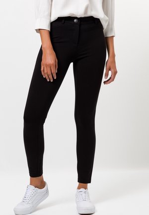 Femme portant un pantalon noir slim, un chemisier blanc à manches longues et des baskets blanches, debout devant un fond blanc uni.