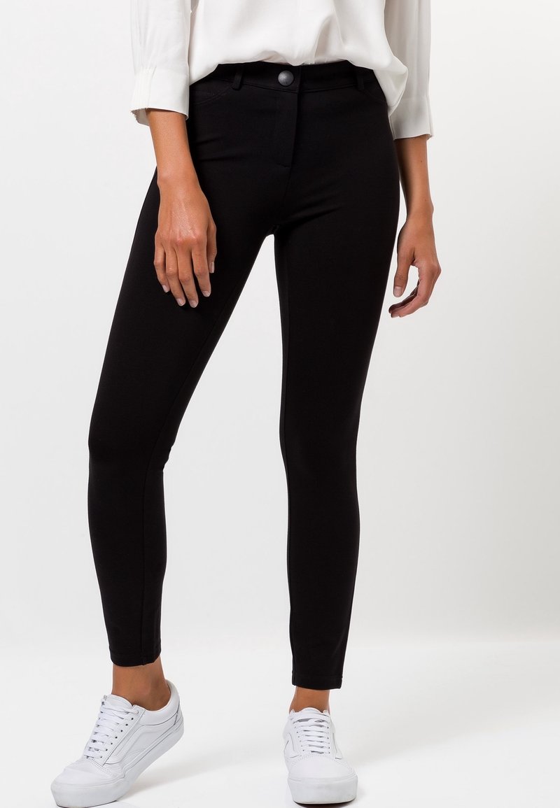 Femme portant un pantalon noir slim, un chemisier blanc à manches longues et des baskets blanches, debout devant un fond blanc uni.