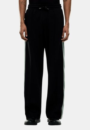 Persona che indossa pantaloni neri a gamba larga con strisce laterali bianche, sneaker nere e top nero, in piedi con le braccia rilassate ai lati.