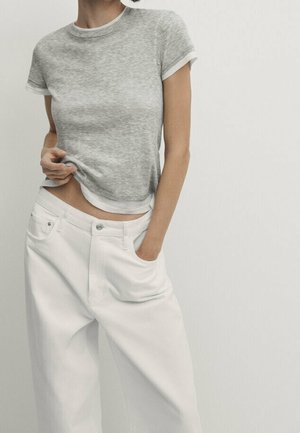 Person wearing a grey short-sleeve shirt and high-waisted loose white pants with one hand in pocket and the other lifting the shirt slightly.