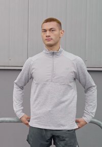 Ljust grå atletisk pullover med kvartdragkedja, kontrasterande textur och långa ärmar. Buren över mörka shorts, visad i en utomhusmiljö.