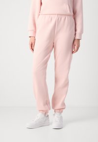 Άτομο ντυμένο με ανοιχτό ροζ παντελόνι και αντίστοιχο φούτερ, σε συνδυασμό με λευκά αθλητικά παπούτσια, σε λευκό φόντο.