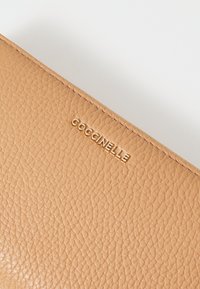 Portafoglio in pelle marrone chiaro con superficie testurizzata e logo "Coccinelle" in rilievo dorato sul fronte.