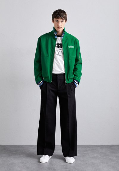 Zelená bunda na zips s modrými prúžkovanými manžetami, navlečená na biele tričko s grafikou, skombinovaná s čiernymi nohavicami širokého strihu a bielymi teniskami.