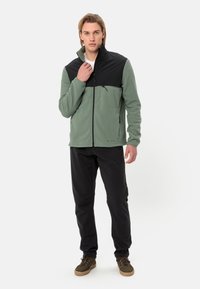Giacca in pile con corpo verde e parte superiore nera, chiusura con zip completa, tasca con zip, indossata con pantaloni neri e scarpe marroni.