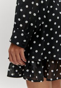 Schwarzes Kleid mit Pünktchen in Weiß, das aus transparentem Stoff gefertigt ist, einen fransigen Saum hat und an den langen Ärmeln über Knopfmanschetten verfügt. Die weißen Punkte heben sich kontrastreich vom Schwarz ab.