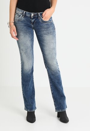 Femme portant un jean bleu délavé droit, des bottines noires à talons et un haut noir, debout avec une main dans la poche.