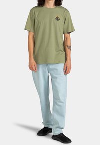 T-shirt en coton vert olive avec un col rond, des manches courtes, et un logo noir et orange sur la poitrine, porté avec un jean bleu clair.