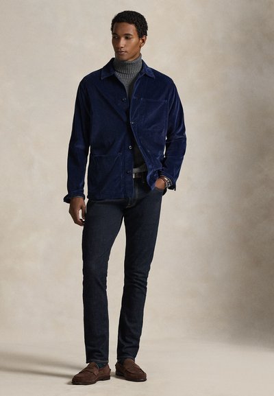 Άνδρας με μπλε σκούρο βελούδινο σακάκι πάνω από γκρι ζιβάγκο, στενά σκούρα τζιν και καφέ σουέτ μοκασίνια, όρθιος με το ένα χέρι στην τσέπη.