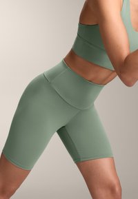 Zelené atletické šortky s vysokým pásom, elastický materiál, hladká textúra, dĺžka do polovice stehna, minimálne švy, vhodné na športové oblečenie.