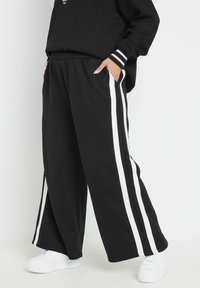 Pantalons de survêtement larges noirs avec des rayures blanches sur les côtés, dotés d'une taille élastique et de poches, associés à des baskets blanches.