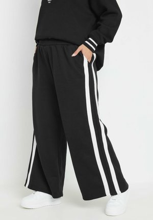 Pantalons de survêtement larges noirs avec des rayures blanches sur les côtés, dotés d'une taille élastique et de poches, associés à des baskets blanches.