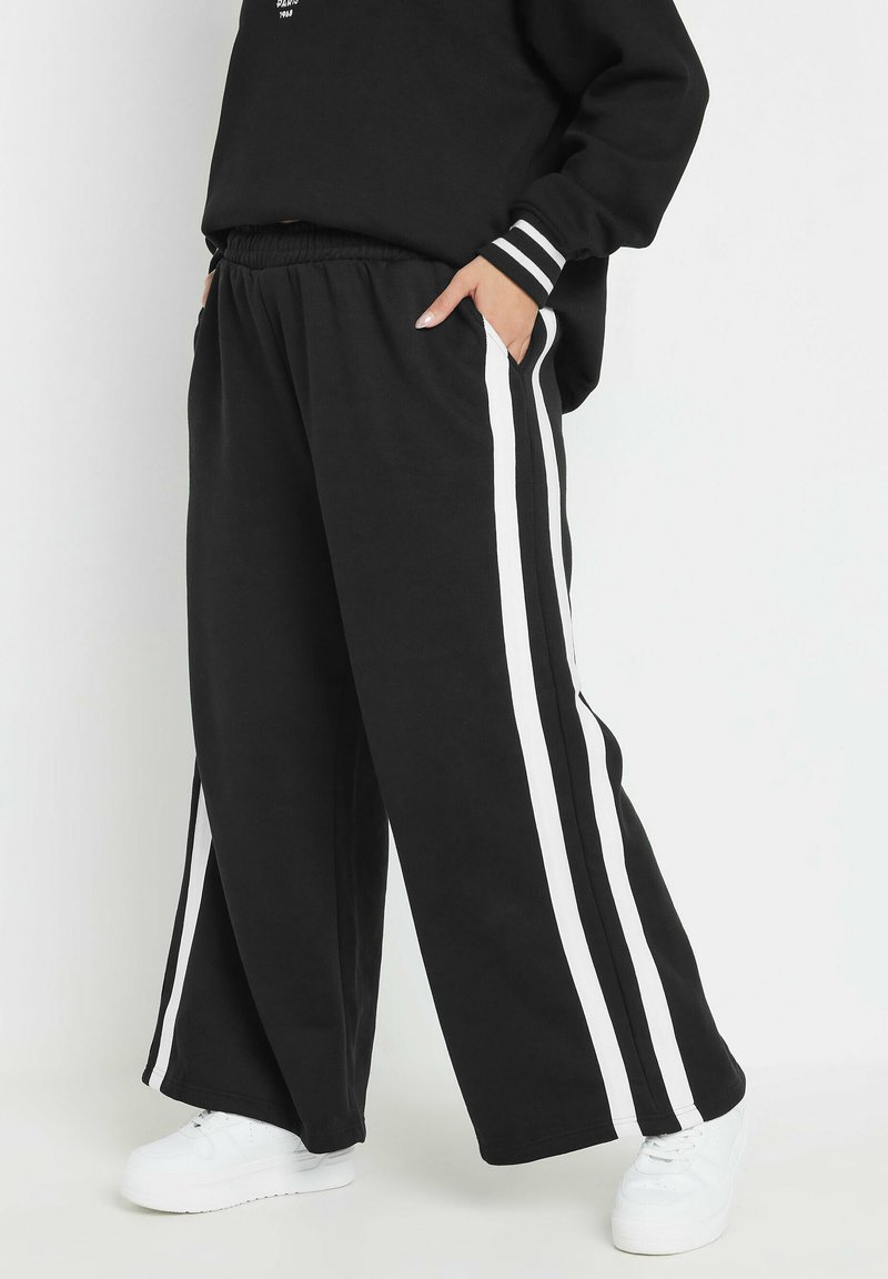 Pantalons de survêtement larges noirs avec des rayures blanches sur les côtés, dotés d'une taille élastique et de poches, associés à des baskets blanches.
