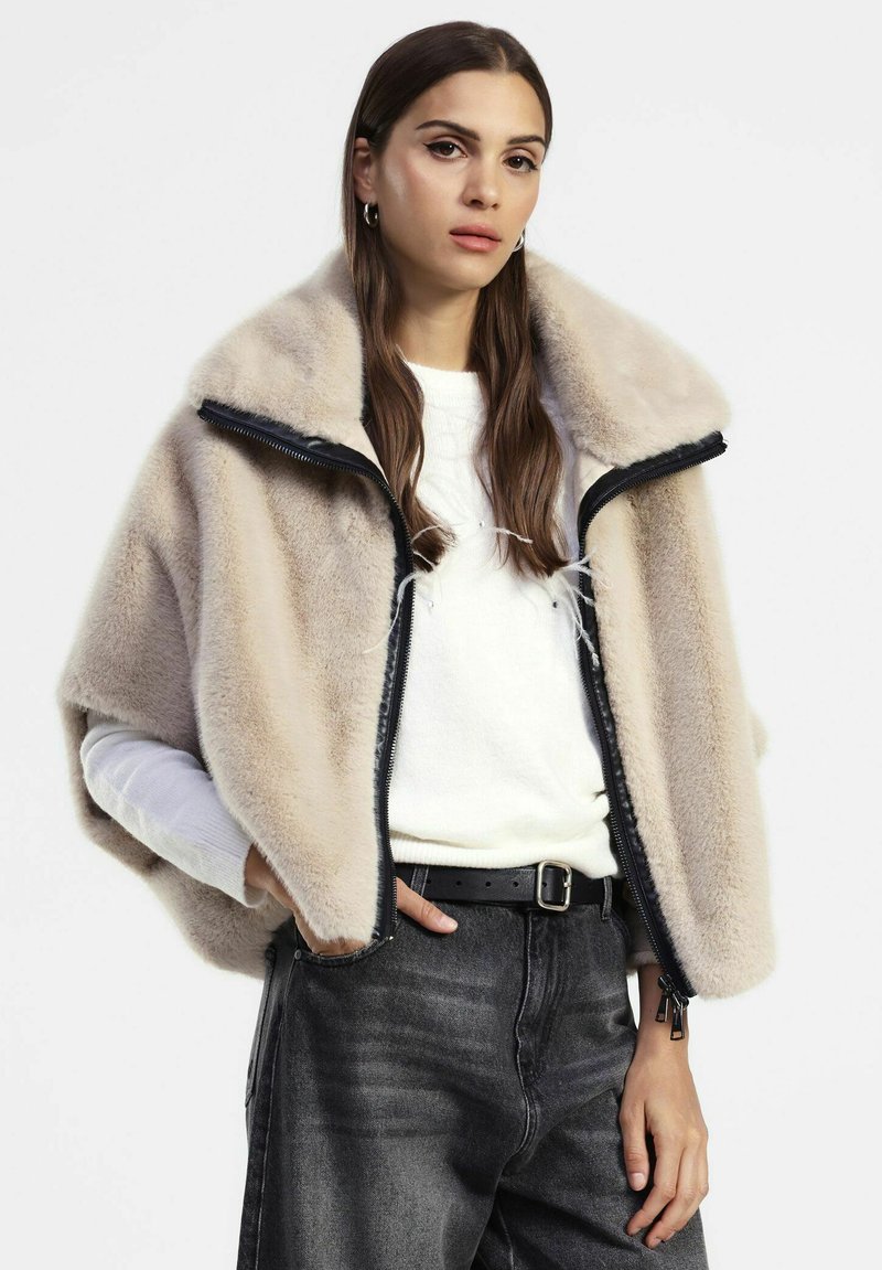 Donna che indossa una giacca in pelliccia sintetica beige sopra un maglione bianco e jeans scuri a vita alta con una cintura nera, in piedi con le mani nelle tasche.
