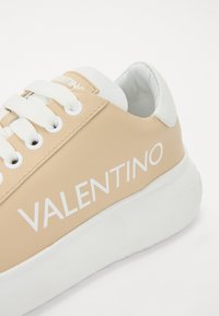 Brune sneakers med hvite detaljer, laget av glatt syntetisk materiale, rund tå, hvite snører og en fremtredende "VALENTINO"-logo på siden.