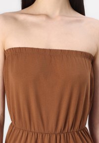 Brązowa sukienka bez ramiączek z zebranym materiałem na górze i elastycznym pasem. Materiał wygląda na gładki i lekki, odpowiedni na ciepłe dni.