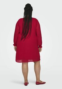 Czerwona sukienka do kolan z teksturowanym wzorem w kropeczki, z długimi rękawami i elastycznymi mankietami. Noszona z dopasowanymi płaskimi butami. Widok z tyłu.