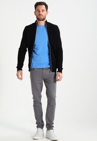 Schwarzer Reißverschluss-Pullover über einem blauen T-Shirt, kombiniert mit grauen Hosen und weißen Sneakers. Der Stoff wirkt glatt; das Design ist figurbetont und lässig.