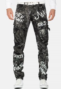 Pantalons cargo noirs avec texte en style graffiti blanc, dotés de poches cargo et de clous décoratifs ; fabriqués à partir d'un tissu texturé avec des effets de taches.