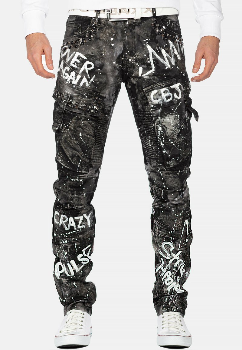 Pantalons cargo noirs avec texte en style graffiti blanc, dotés de poches cargo et de clous décoratifs ; fabriqués à partir d'un tissu texturé avec des effets de taches.