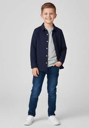 Chłopiec ubrany w granatową koszulę na guziki, szarą koszulkę, niebieskie dżinsy i białe trampki, stojący swobodnie i uśmiechnięty.