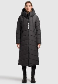 Long black puffer coat with chevron pattern, hood, and zippered pockets; features a logo tag on the front and is paired with black boots.