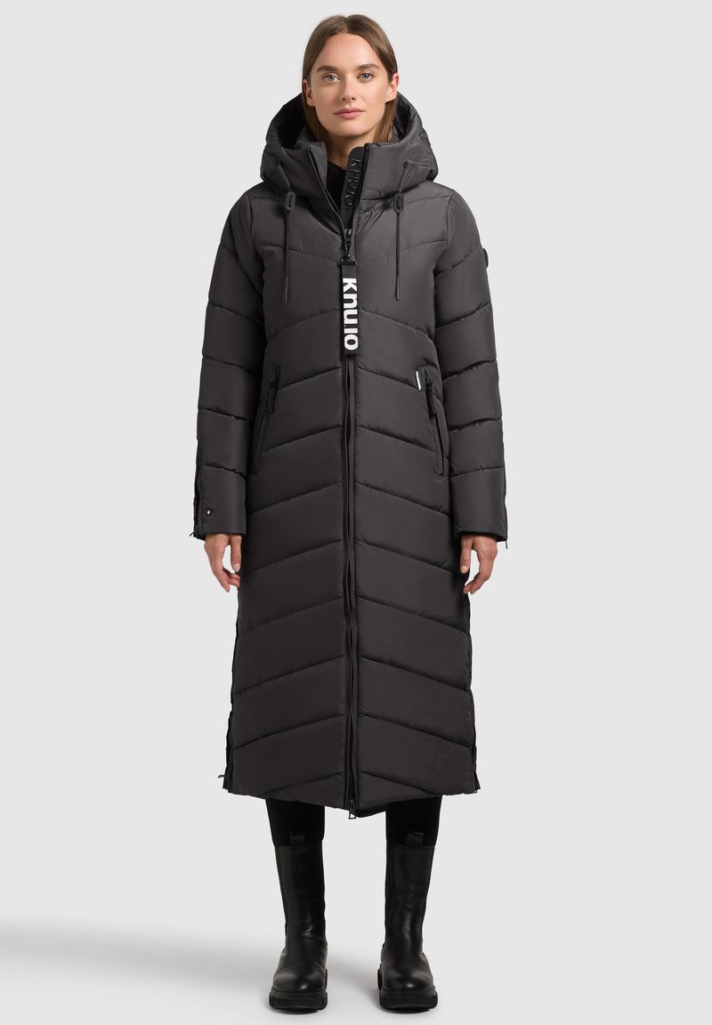 Long black puffer coat with chevron pattern, hood, and zippered pockets; features a logo tag on the front and is paired with black boots.