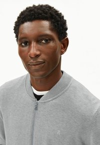 Graue Bomberjacke mit Reißverschluss und geripptem Kragen. Verfügt über ein kleines Logo auf der Brust und ein weißes Innenfutter. Glatte Stoffstruktur.