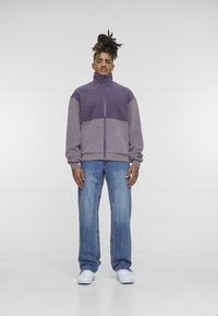 Chaqueta de forro polar morada con cuello alto, cierre de cremallera y dos bolsillos, combinada con unos vaqueros de mezclilla azul claro y zapatillas blancas. Diseño simple.