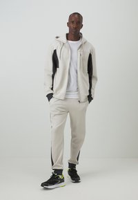 Sudadera gris claro y joggers a juego con paneles laterales negros. Camiseta blanca debajo. Zapatillas negras con acentos en amarillo neón.