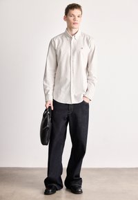 Πουκάμισο με λευκές και μπεζ ρίγες και κουμπιά, μακριά μανίκια, συνδυασμένο με σκούρα μπλε παντελόνι με φαρδιά μπατζάκια· κρατώντας μια μαύρη δερμάτινη τσάντα.